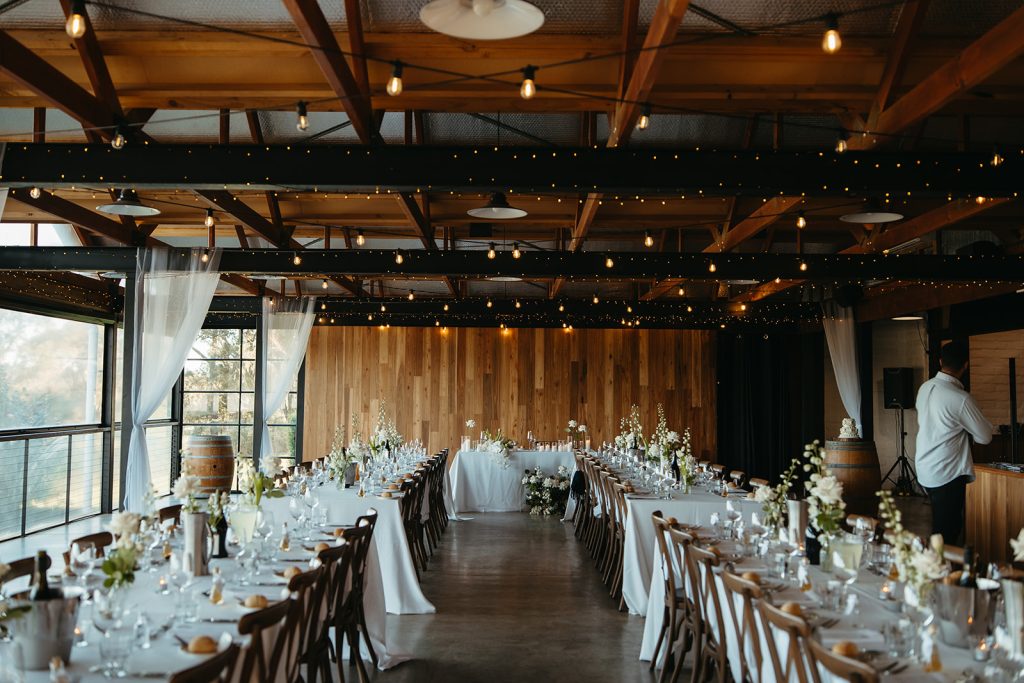 Romantic summer wedding reception at Yarra Ranges Estate with festoon lighting, a timber wall backdrop and a vibrant summery atmosphere with flowers & candles on reception tables.